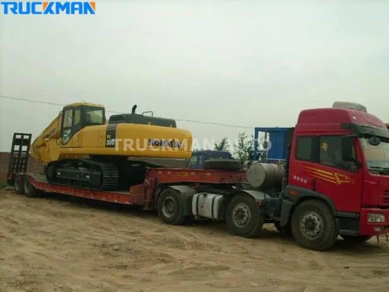 Heavy yellow excavator being transported on a red low-bed semi-trailer on a dirt road