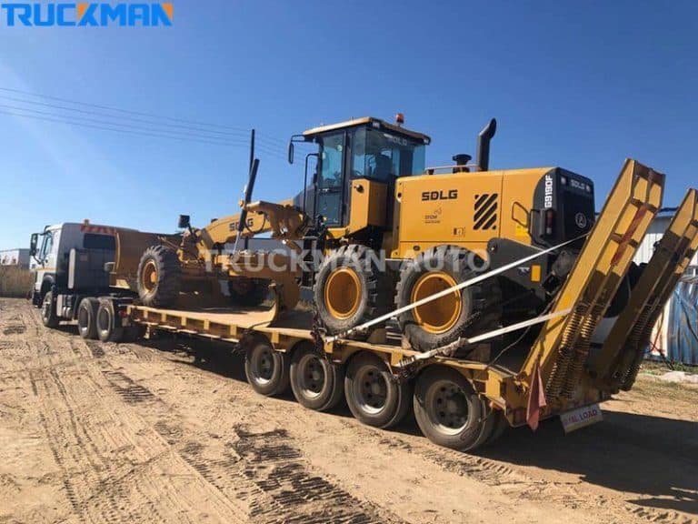 Large construction grader secured on a multi-axle low-bed semi-trailer in a dirt lot