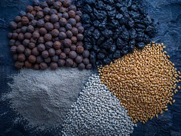 Vue rapprochée de cinq échantillons secs en vrac — minerai de fer, charbon, céréales, poudre de ciment et phosphate — disposés sur une surface industrielle sombre